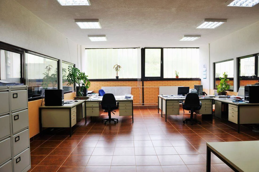 Modern office space with desks, black chairs, plants, and large windows with brick accent wall