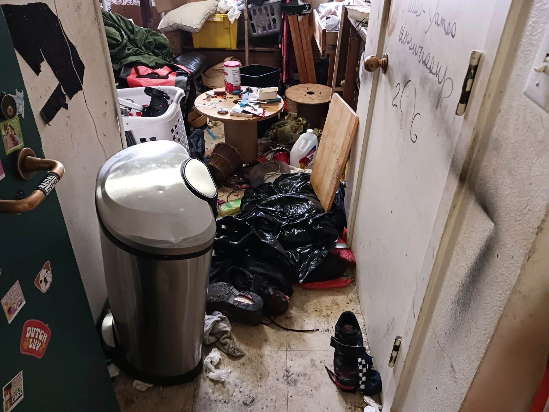 Cluttered room with trash can, scattered clothing, and household items on floor