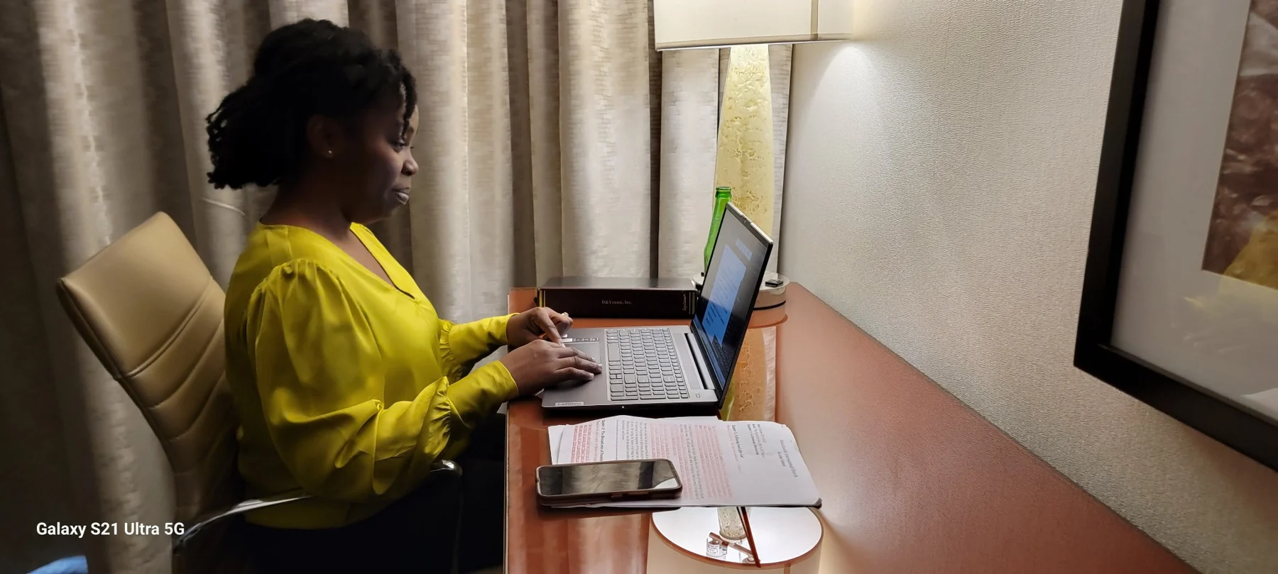 Woman in yellow shirt working at laptop at wooden desk with documents and phone