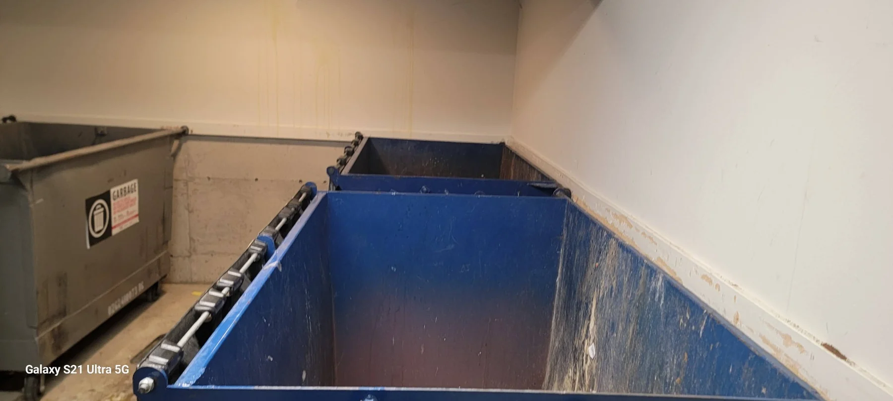 Large blue industrial waste bin in storage room with beige walls and brown container on left