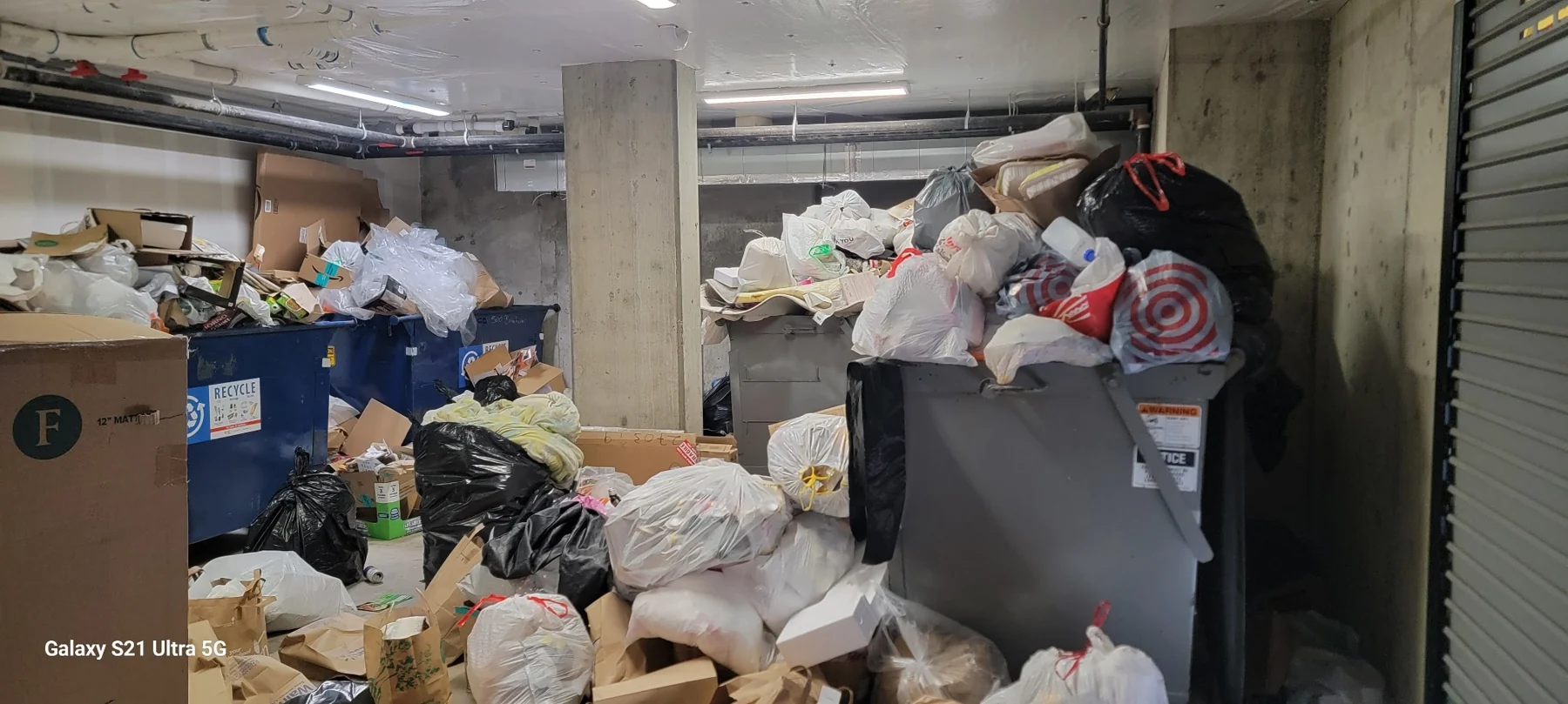 Cluttered basement storage area with overflowing trash bins, cardboard boxes, and plastic bags stacked against concrete pillars