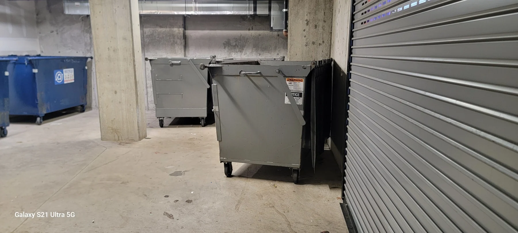 Dumpsters and recycling bins in underground parking garage next to metal roll-up door